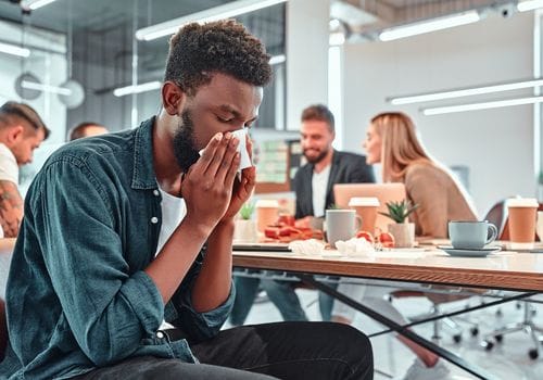 Unwell office worker blowing his nose at a meeting, needing a medical certificate for sick leave