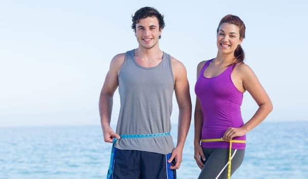 Man and woman measuring their waists on a Gold Coast beach, representing GP-led weight management at My Doctors Clinic Surfers Paradise